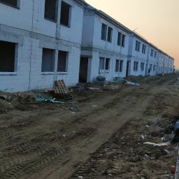 RASBUD SPÓŁKA Z OGRANICZONĄ ODPOWIEDZIALNOŚCIĄ - Wznoszone budynki bliźniacze z białych bloczków, widoczne otwory okienne i drzwiowe. Teren wokół w stanie surowym, przygotowany pod dalsze prace budowlane, w oddali zachodzące słońce.