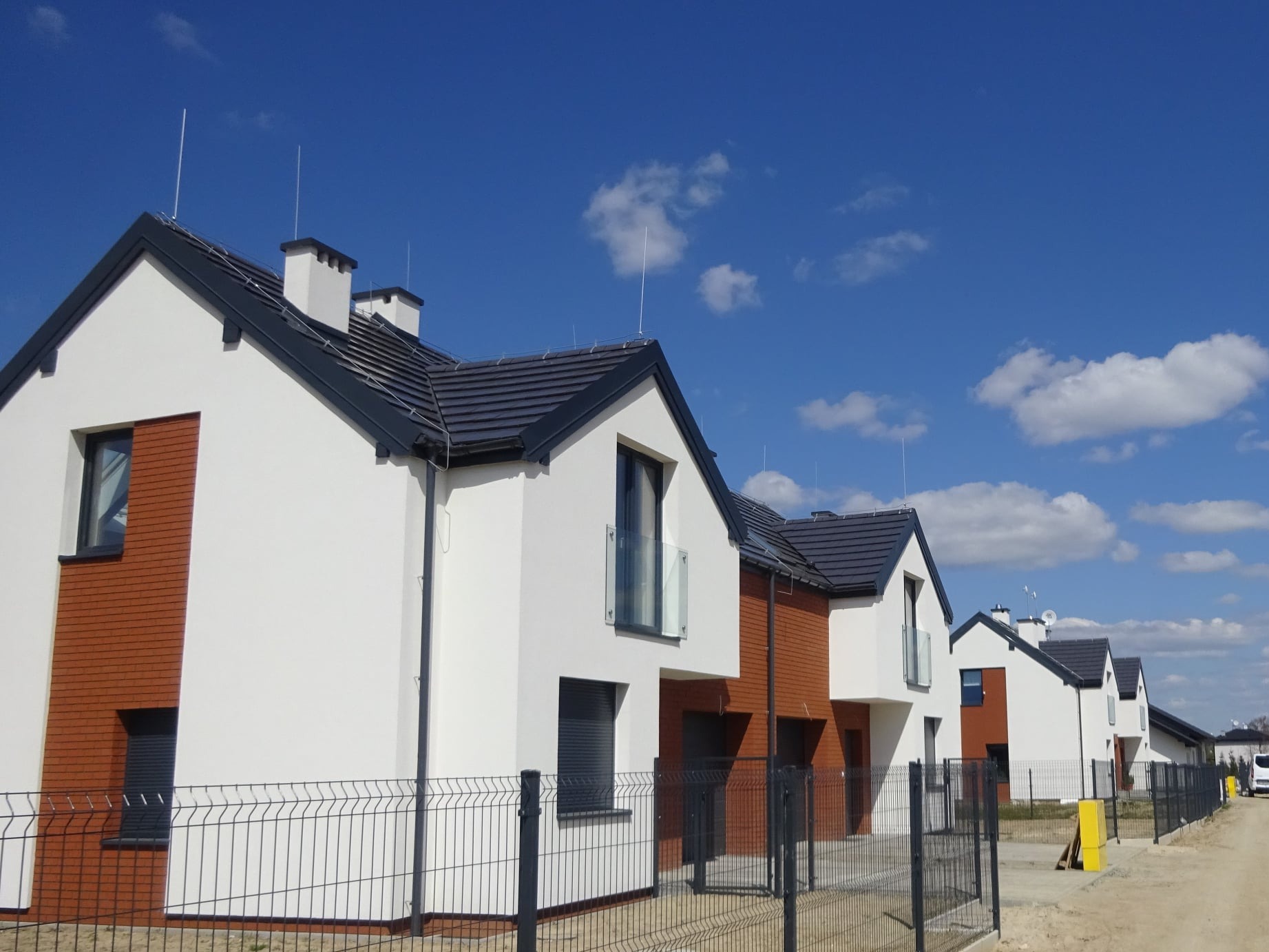 Szereg nowych domów jednorodzinnych w stanie deweloperskim, z białymi elewacjami i elementami z cegły, ciemnymi dachami i ogrodzeniem panelowym na tle błękitnego nieba.