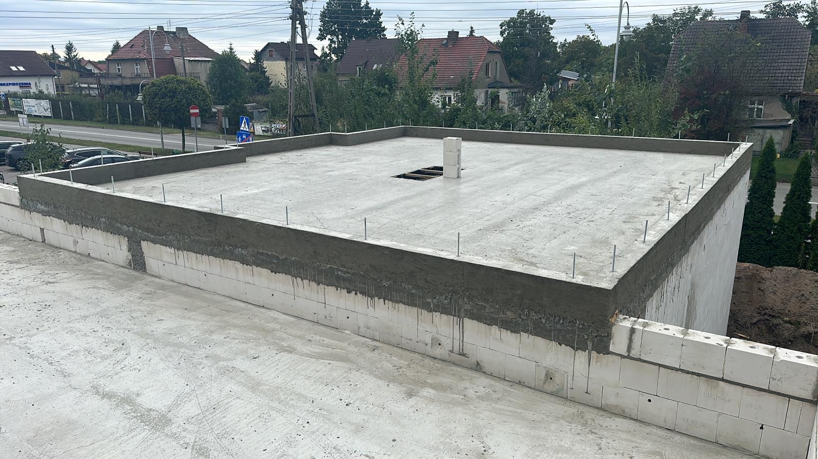 Budowa budynku w stanie deweloperskim: ściany z betonu komórkowego, wylany strop, wystające pręty zbrojeniowe. Widok z góry na plac budowy i okoliczną zabudowę.