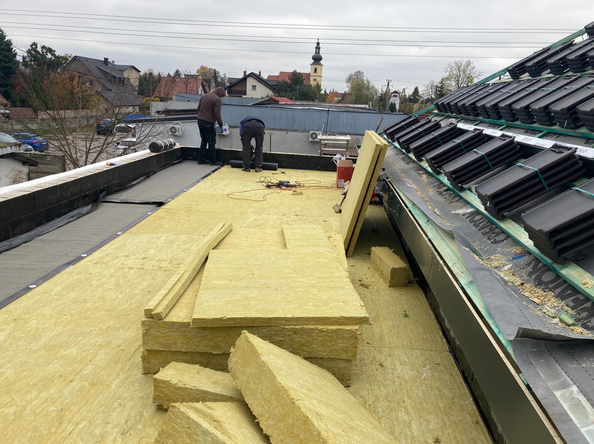 Montaż izolacji z wełny mineralnej na płaskim dachu budynku z częściowo ułożoną dachówką. Dwóch pracowników przy pracy, widok na okoliczne budynki i kościół.