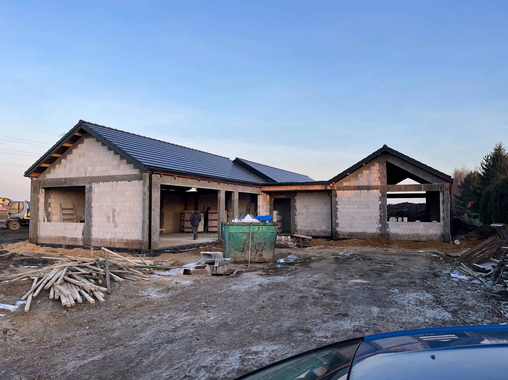 Budowa domu jednorodzinnego w stanie surowym otwartym: ściany z białego bloczka, dach pokryty ciemną dachówką, widoczny kontener na gruz i materiały budowlane.