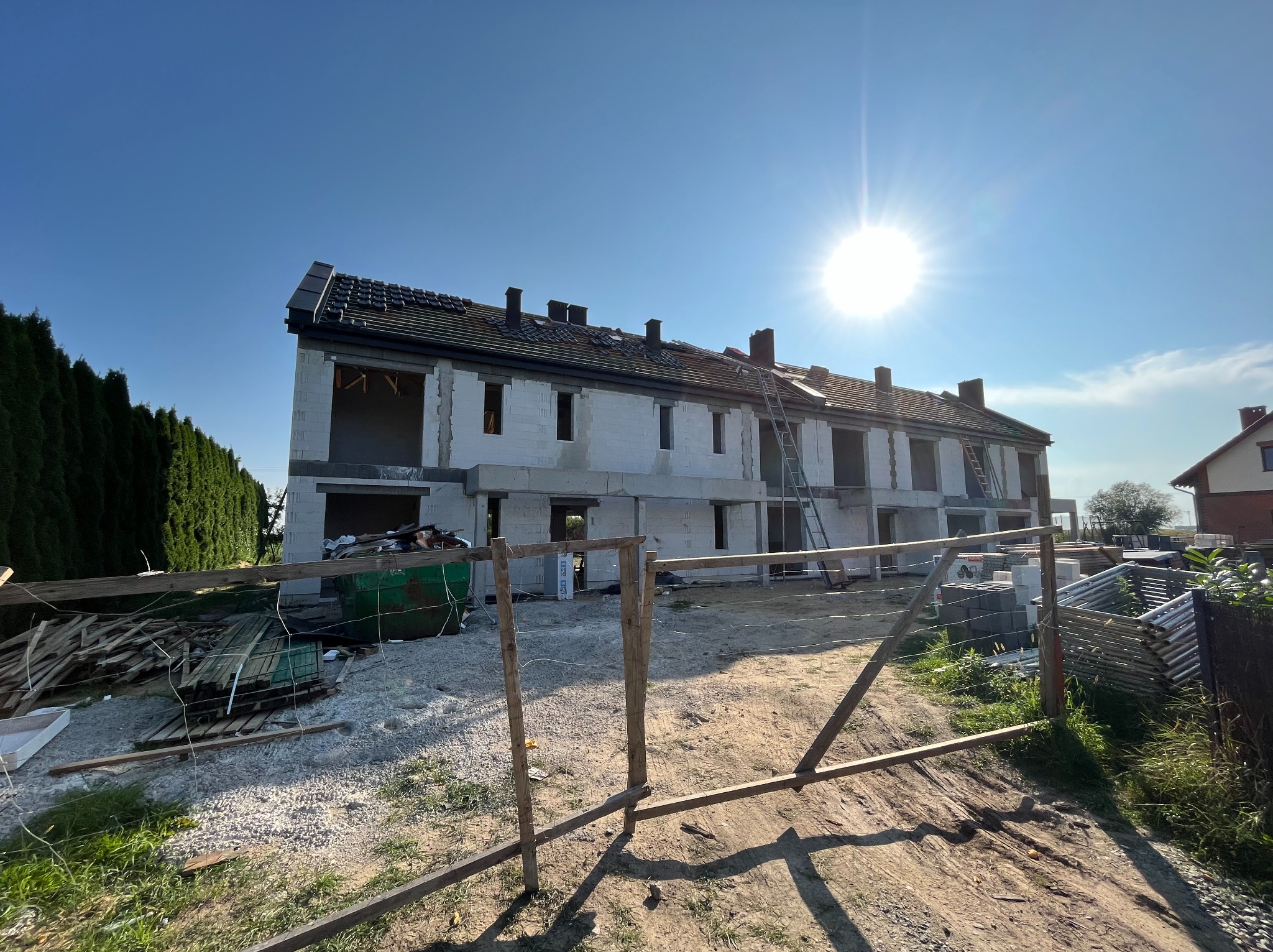 Budynek bliźniaczy w stanie deweloperskim, z częściowo wykonanym dachem i widocznymi pustakami. Na pierwszym planie prowizoryczne ogrodzenie i materiały budowlane.