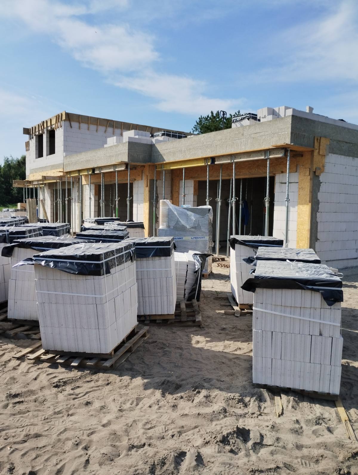 Budowa domu jednorodzinnego w Dziwnowie. Widoczne ściany z białych bloczków, podpory stropu i palety z materiałami budowlanymi. Częściowo zrealizowany etap murowania ścian.