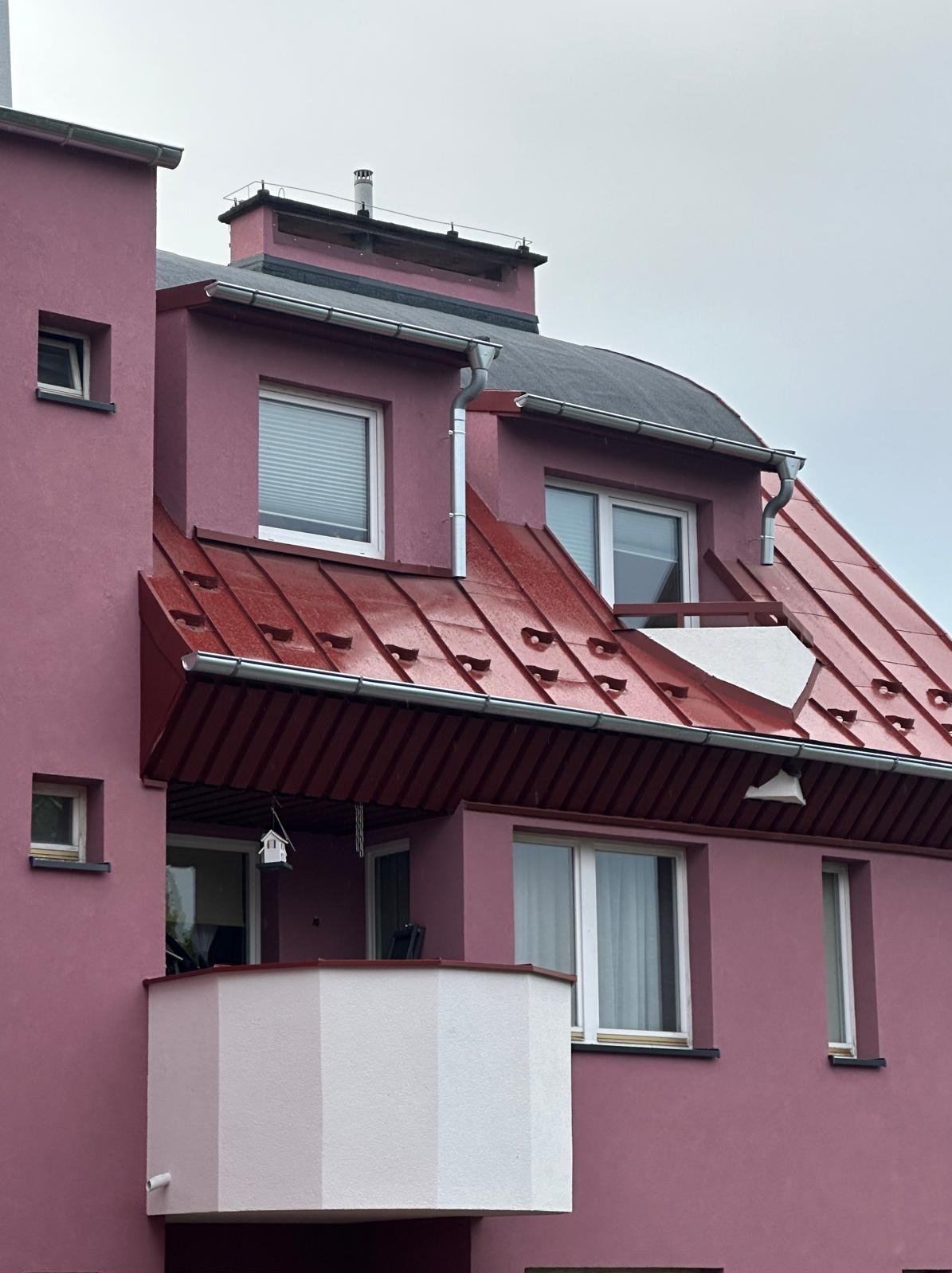 Różowa elewacja budynku z czerwonym dachem z blachy trapezowej, widoczne okna z roletami i balkon z białą balustradą. Na dachu komin.