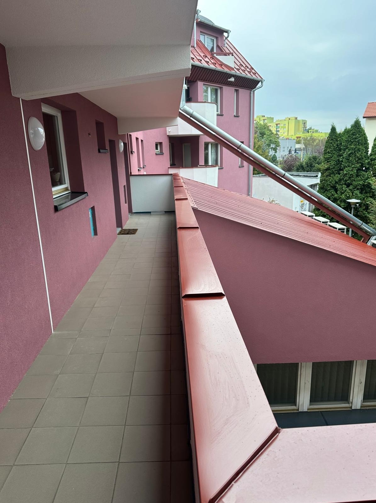Odnowiona elewacja i dach budynku wielorodzinnego w odcieniach różu, widok z zewnątrz z poziomu balkonu. Metalowa rynna biegnie wzdłuż ściany.