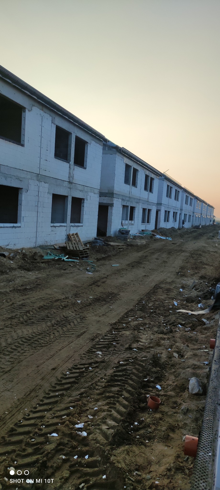 Wznoszone budynki bliźniacze z białych bloczków, widoczne otwory okienne i drzwiowe. Teren wokół w stanie surowym, przygotowany pod dalsze prace budowlane, w oddali zachodzące słońce.