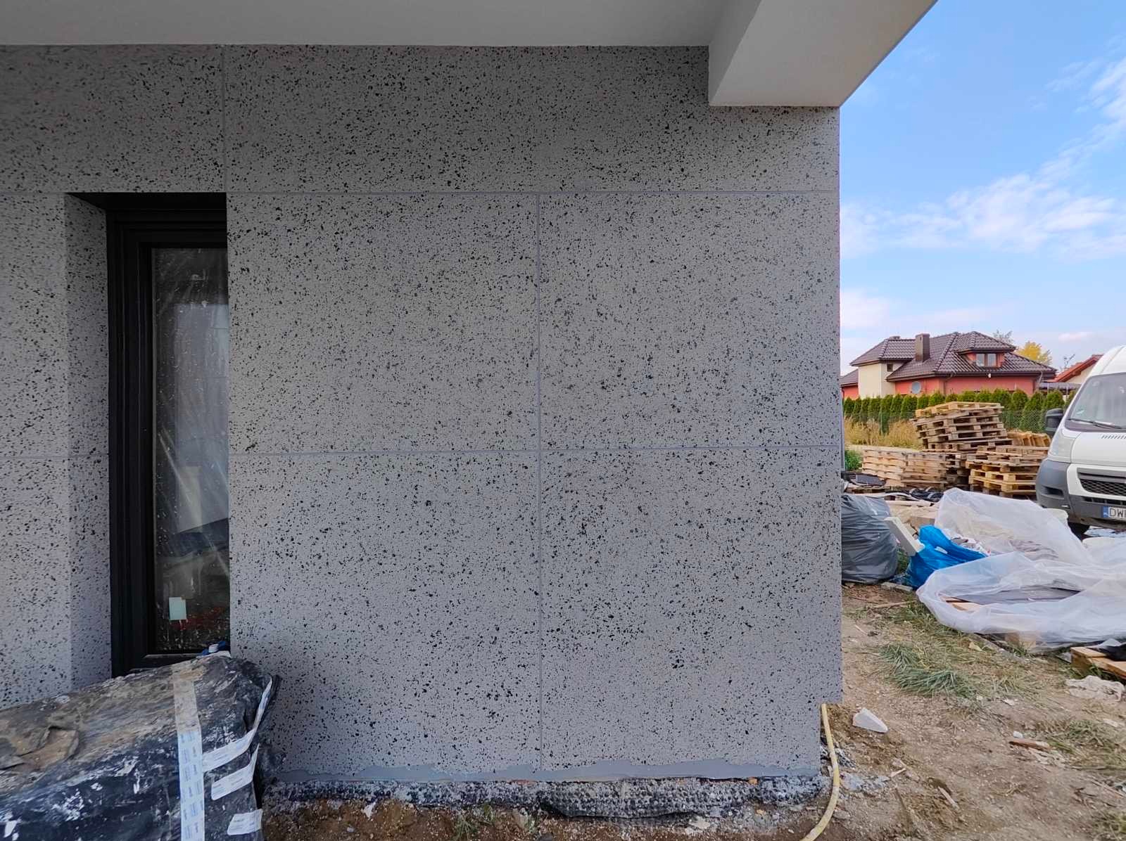 Szara elewacja domu jednorodzinnego z oknem, wykończona panelami z fakturą. W tle widoczny fragment innego budynku i niebo.