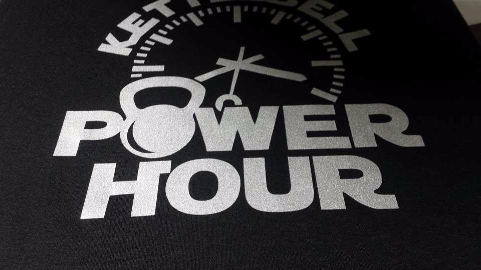 Czarna koszulka z srebrnym nadrukiem 'KETTLEBELL POWER HOUR' i grafiką kettlebell w formie tarczy zegara.