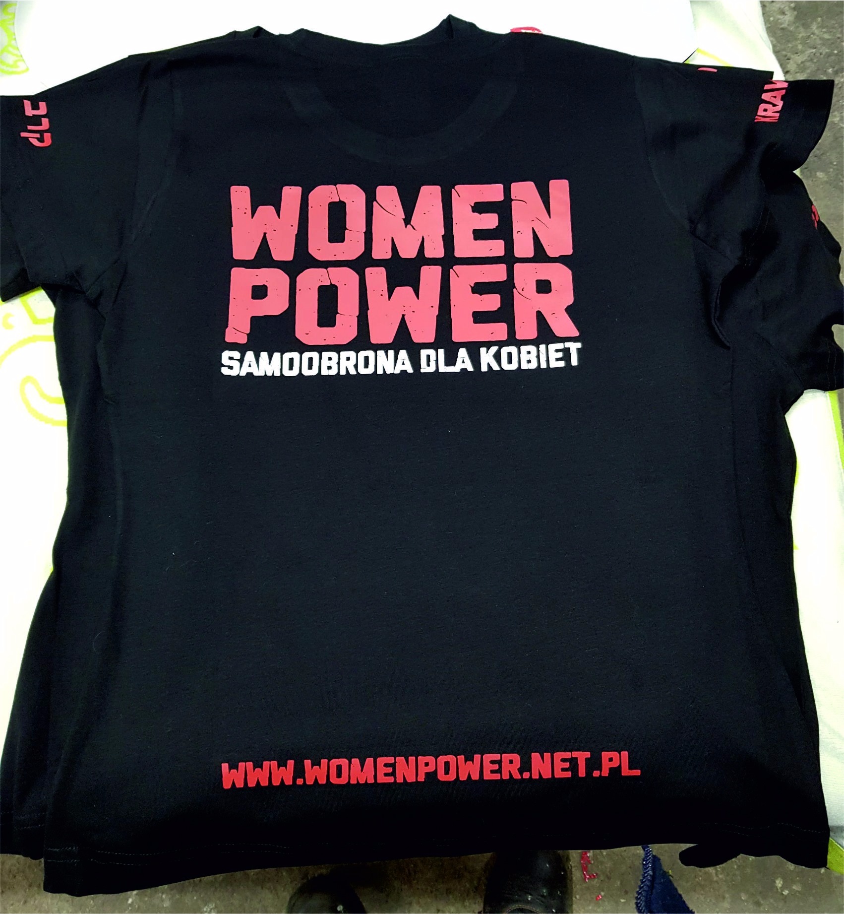 Czarna koszulka z różowym nadrukiem 'WOMEN POWER SAMOOBRONA DLA KOBIET' i adresem strony internetowej na dole.