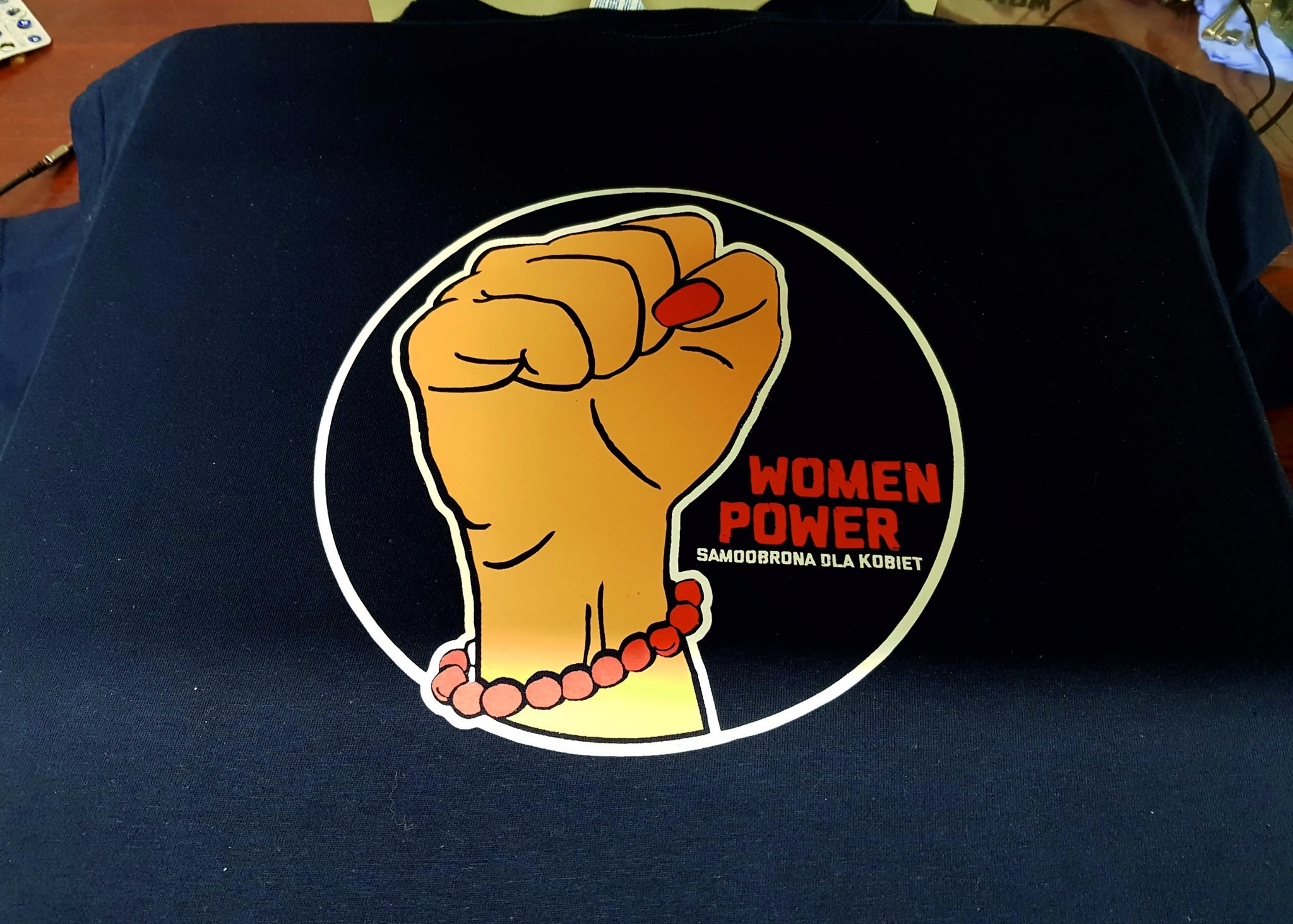 Granatowa koszulka z nadrukiem pięści w okręgu, z napisem 'WOMEN POWER SAMOOBRONA DLA KOBIET', widoczny fragment rękawa i górnej części koszulki.