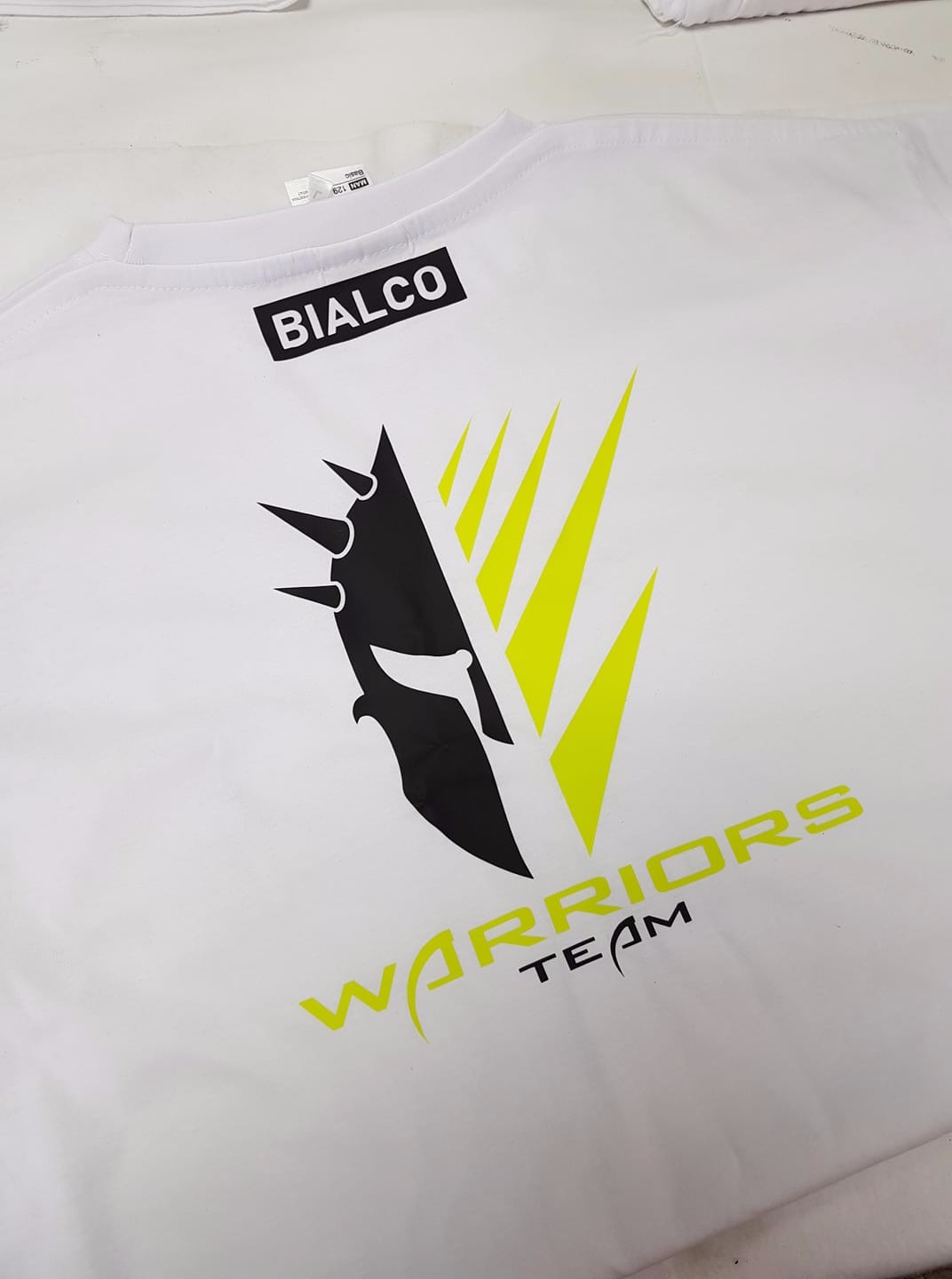 Biała koszulka z nadrukowanym czarno-żółtym logo przedstawiającym hełm wojownika i napis 'WARRIORS TEAM' oraz nazwą firmy 'BIALCO'.