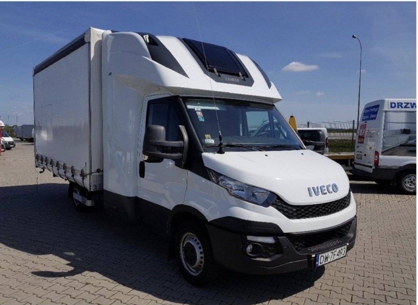 Biały samochód dostawczy marki Iveco z plandeką na parkingu, widok z przodu i boku.