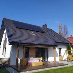 Nowoczesny dom jednorodzinny z brązową dachówką, panelami fotowoltaicznymi na dachu i drewnianym tarasem z meblami ogrodowymi w słoneczny dzień.
