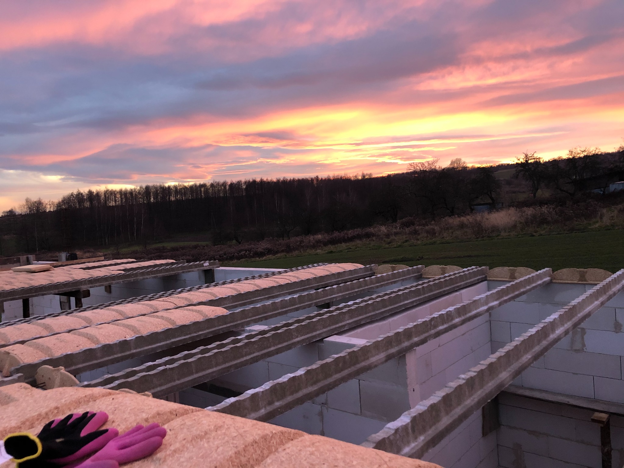 Wznoszenie fundamentów domu z bloczków betonowych, widoczne zbrojenie, w tle zachodzące słońce barwiące niebo na różowo-pomarańczowo, w rogu kadru leżą robocze rękawice.