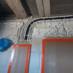 Instalacja elektryczna w surowym wnętrzu: liczne kable i peszle ułożone równolegle na ścianie z betonu komórkowego pod sufitem, zabezpieczone kartonem z pomarańczową taśmą, niebieska folia ochronna...