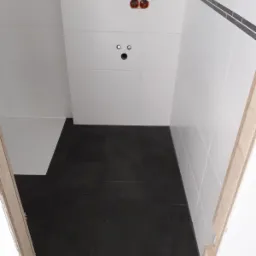 Wykończona łazienka w Łowiczu z czarną podłogą i białymi płytkami ściennymi z ciemnym paskiem dekoracyjnym, widoczne gniazdka elektryczne i wyprowadzenia hydrauliczne.