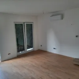 Jasne wnętrze po remoncie z ułożonymi panelami podłogowymi, widoczne okno balkonowe z roletami, klimatyzator na ścianie, gładkie ściany przygotowane do malowania, oraz otwór drzwiowy bez ościeżnicy...