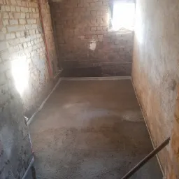 Pomieszczenie w trakcie prac remontowych, widoczne surowe, ceglane ściany, świeżo wylana wylewka podłogowa, narzędzia i butelki z wodą w rogu.