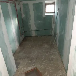 Surowe wnętrze w trakcie prac, ściany wyłożone płytami gipsowo-kartonowymi z widocznymi łączeniami, fragment instalacji grzewczej, surowy beton na podłodze i kartonowe pudło.