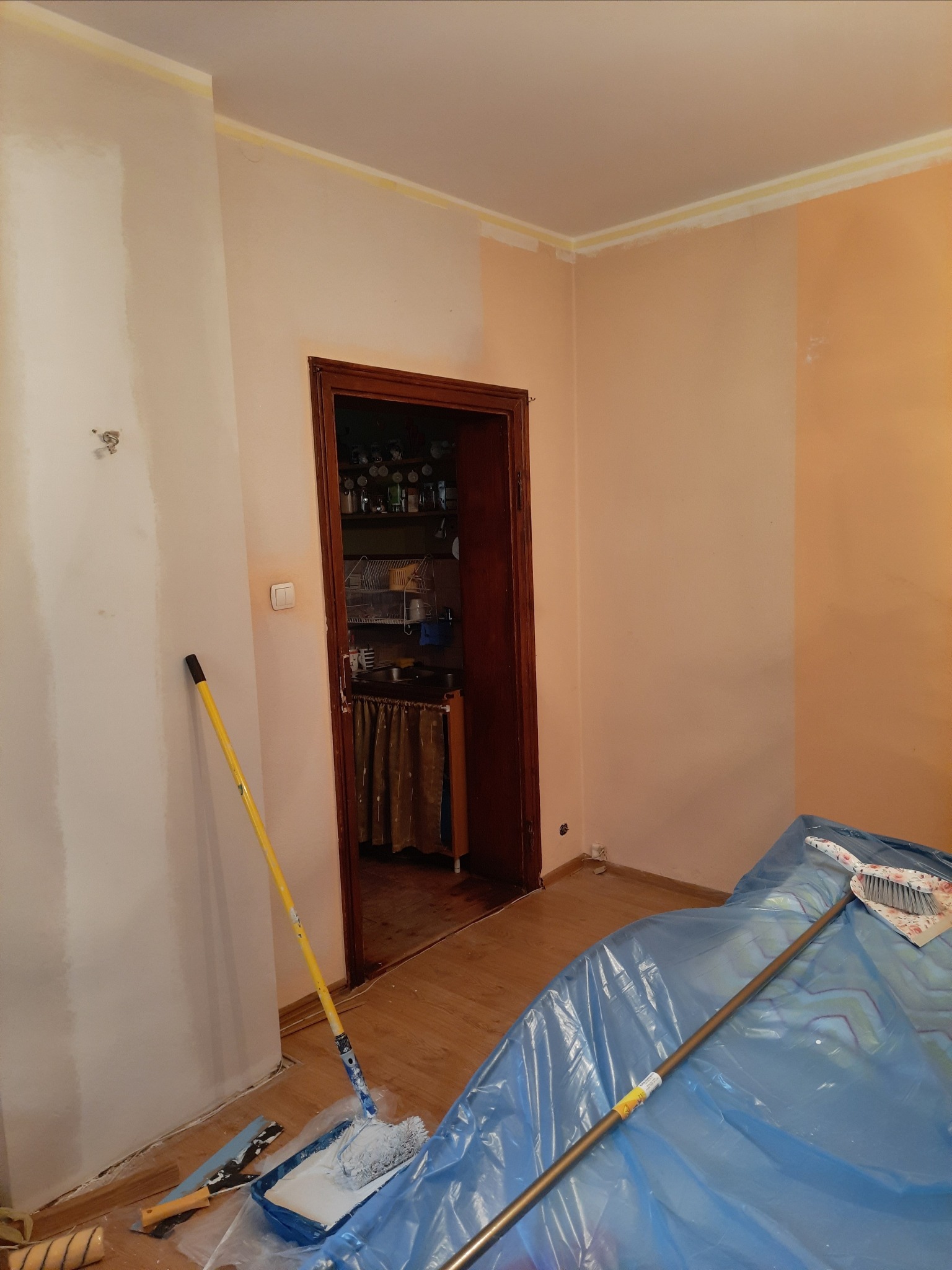 Pokój w trakcie odświeżania: fragment ściany pomalowany na biało, reszta w odcieniach pomarańczy, podłoga zabezpieczona folią, widoczny wałek malarski w kuwecie i kij od szczotki z szufelką.