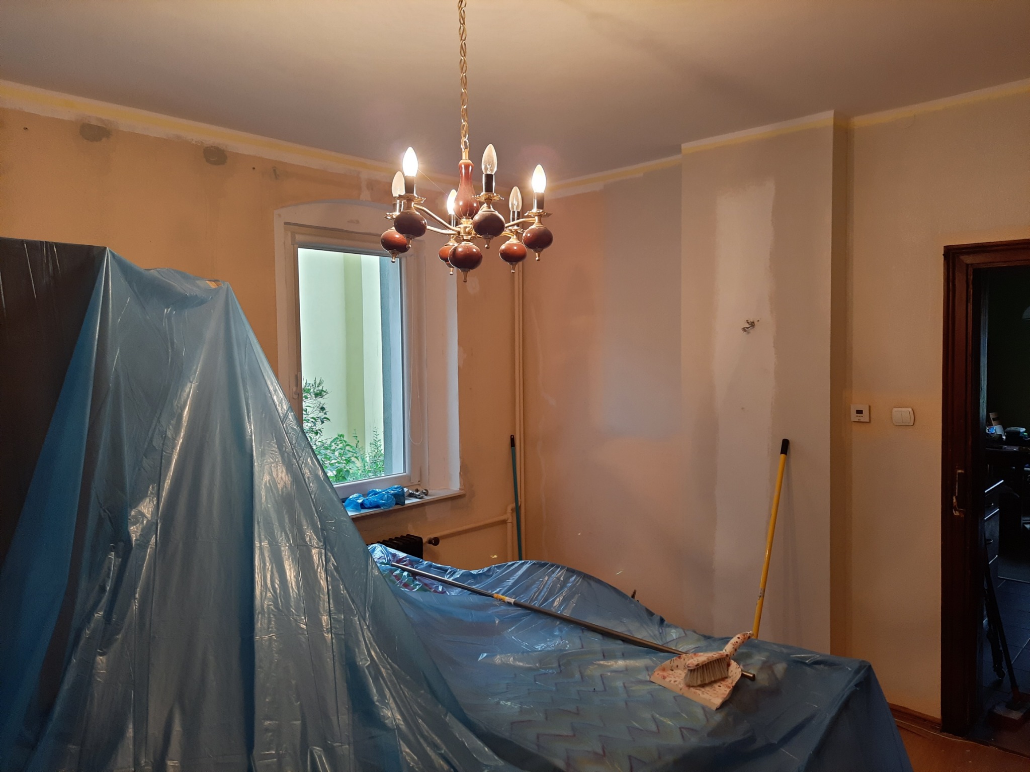 Pokój w trakcie przygotowań do prac wykończeniowych: ściany częściowo zagruntowane, podłoga i meble zabezpieczone folią ochronną, widoczna lampa sufitowa, okno i szczotka z szufelką.