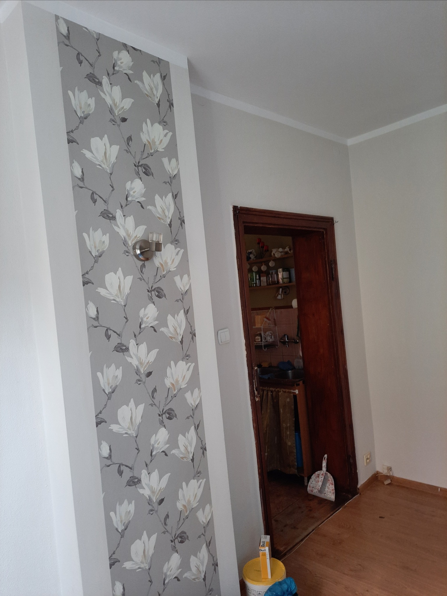 Fragment ściany z szarą tapetą w białe magnolie, oświetlony kinkietem, z widocznym wejściem do kuchni w tle.