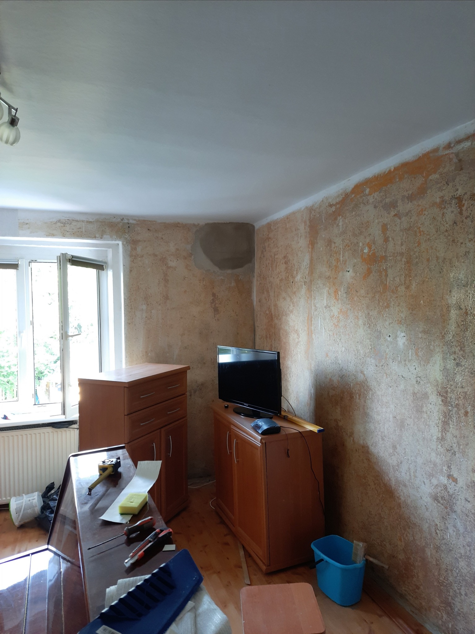 Pokój w trakcie przygotowań do remontu: ściany z resztkami starej farby i zagruntowanymi plamami, meble odsunięte od ścian, narzędzia na blacie stołu.