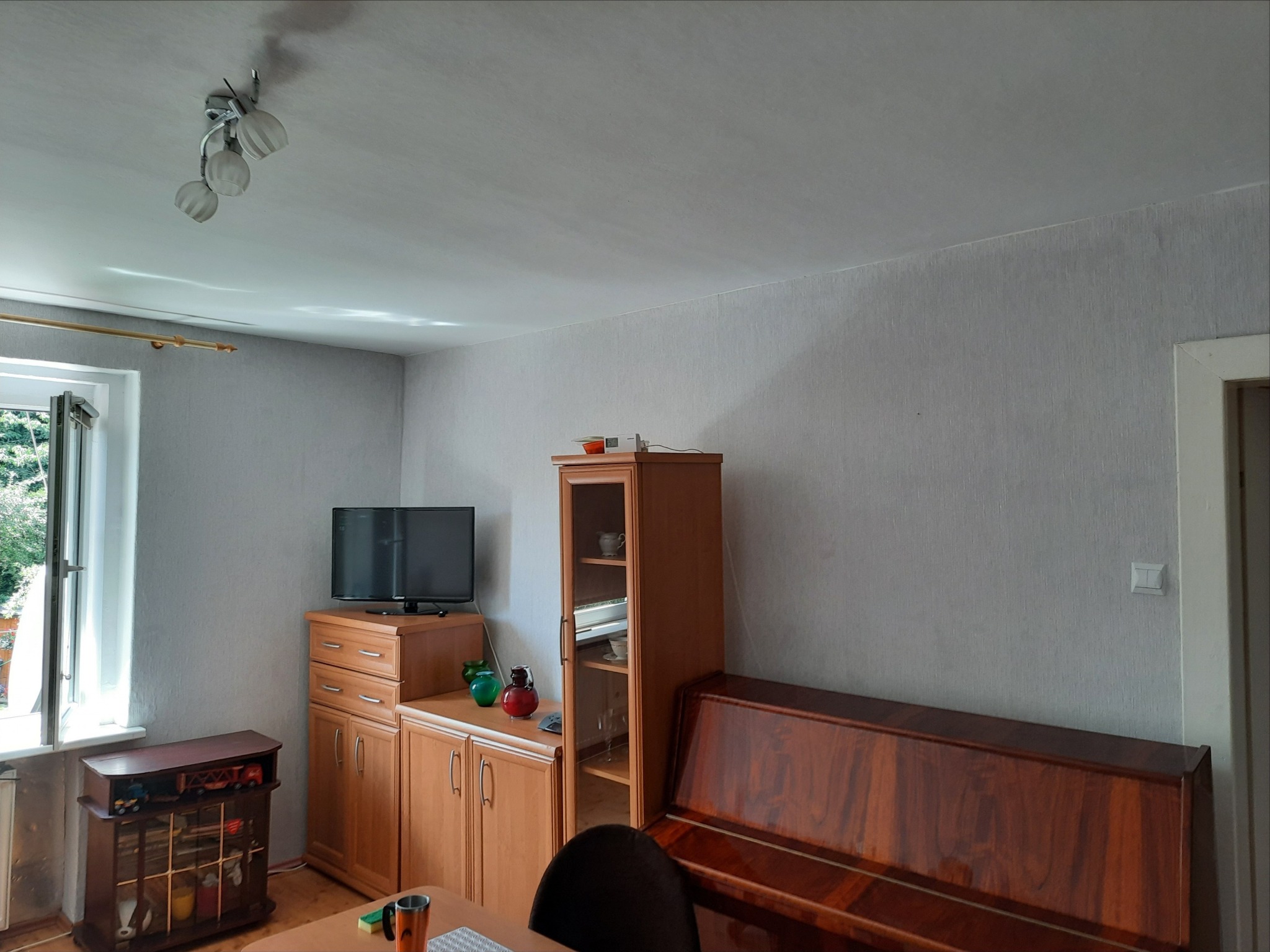 Szara ściana w pokoju po tapetowaniu z widoczną strukturą tapety, oświetlenie sufitowe z trzema kloszami, drewniane meble w tle i okno po lewej stronie.