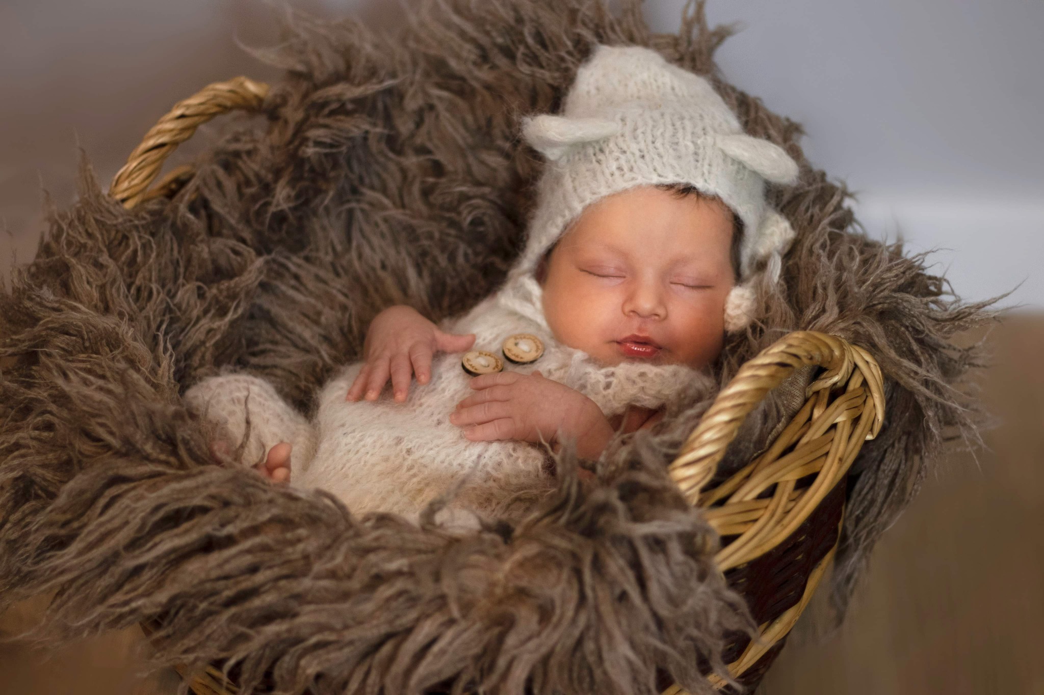 Śpiący noworodek w wełnianym kombinezonie z drewnianymi guzikami i czapce z uszkami, ułożony w koszyku wyłożonym sztucznym futrem.