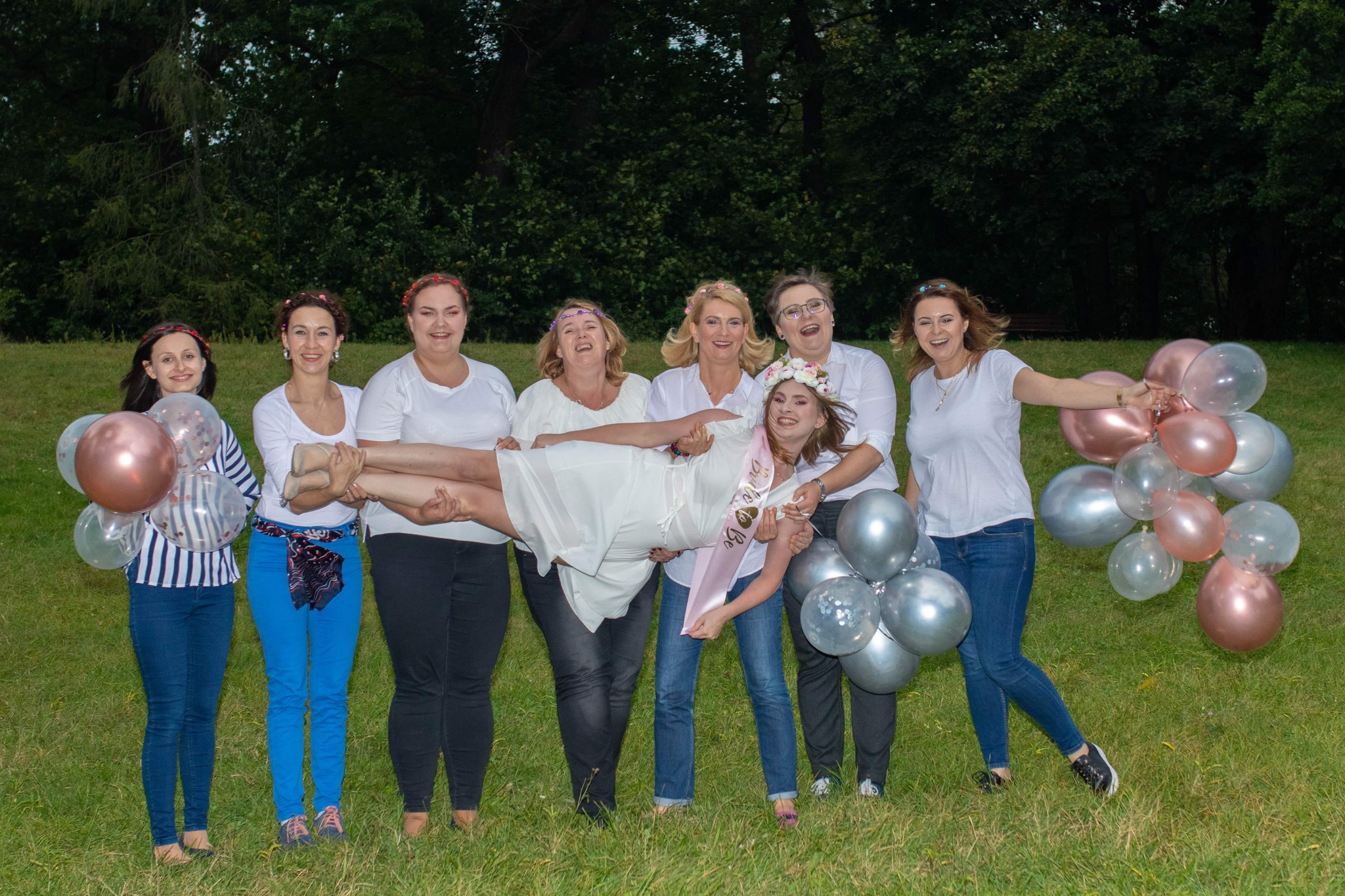 Grupa kobiet świętuje wieczór panieński w plenerze, trzymając przyszłą pannę młodą w powietrzu, w tle zielona trawa i drzewa, a w rękach balony w odcieniach różu i srebra.