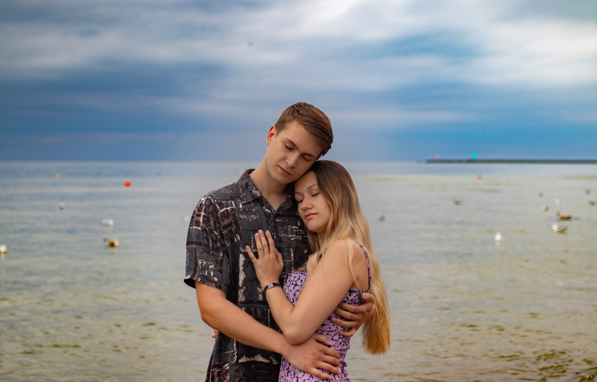 Zakochana para przytulona na plaży z widokiem na morze i pochmurne niebo, kobieta w kwiecistej sukience, mężczyzna w wzorzystej koszuli.