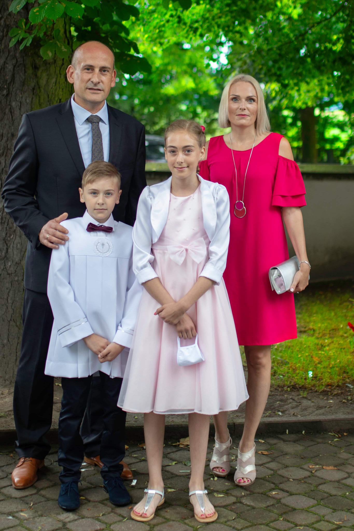 Rodzinne zdjęcie portretowe: ojciec w garniturze obejmuje syna w albie, córka w różowej sukience trzyma maseczkę, matka w fuksjowej sukience z srebrną torebką.