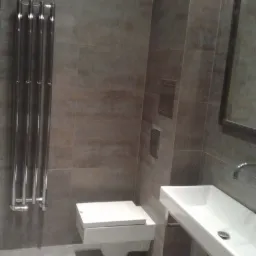 Nowoczesna łazienka w odcieniach szarości z podwieszaną toaletą, prostokątną umywalką i grzejnikiem drabinkowym z chromowanymi rurami.