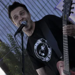 Wokalista z gitarą Gibson śpiewa do mikrofonu na scenie, ujęcie z dołu, widoczny wzór na koszulce.