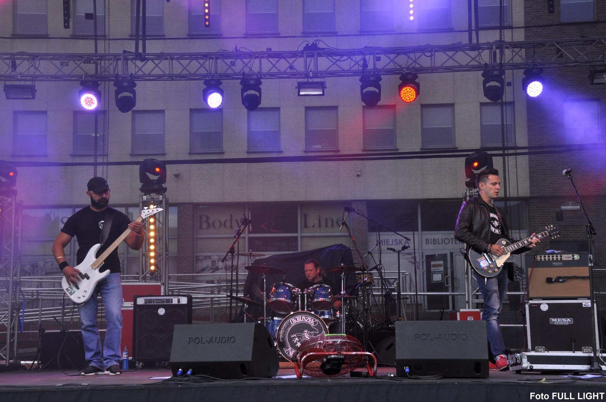 Zespół muzyczny grający na scenie plenerowej, widoczni basista z białą gitarą basową, gitarzysta w skórzanej kurtce grający na gitarze elektrycznej oraz perkusista za bębnami, w tle budynek...