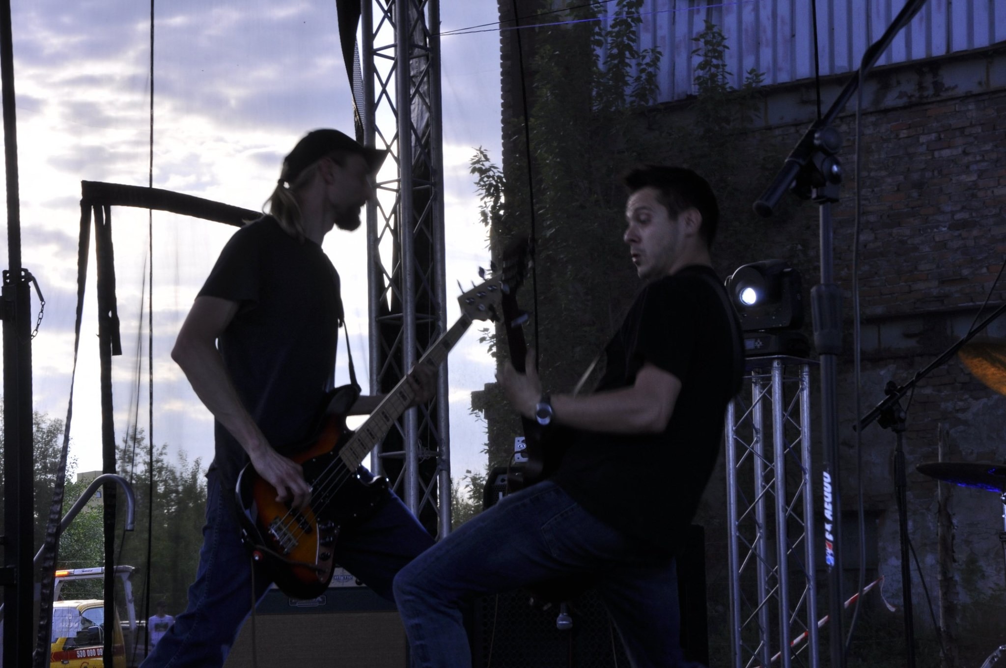 Dwóch basistów gra na scenie; jeden z długimi włosami i czapce, drugi z krótkimi; w tle ceglana ściana i sprzęt oświetleniowy.