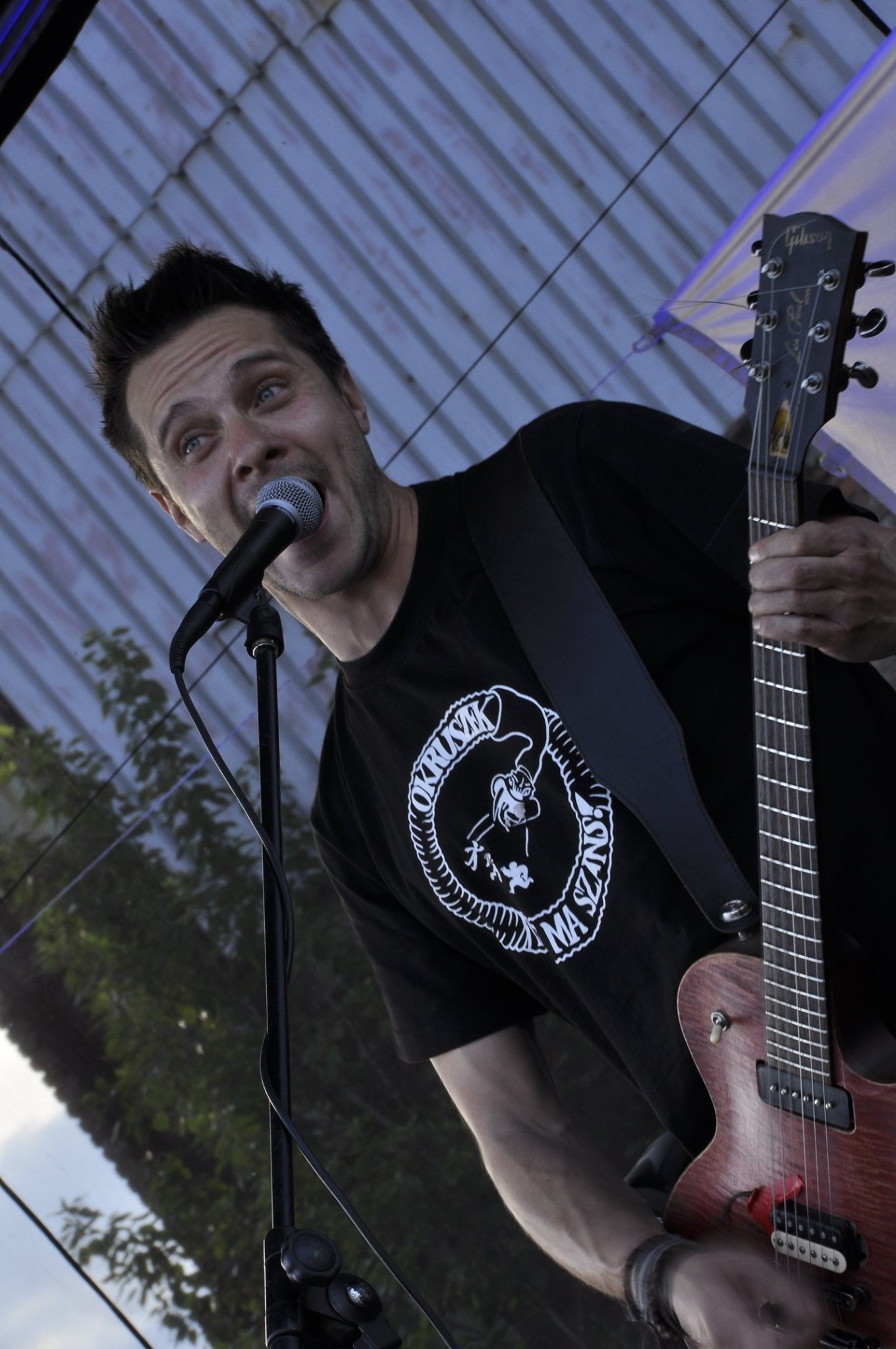 Wokalista z gitarą Gibson śpiewa do mikrofonu na scenie, ujęcie z dołu, widoczny wzór na koszulce.