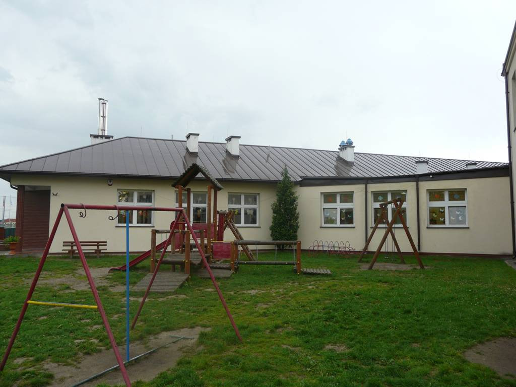 Dach budynku przedszkola pokryty blachą trapezową w kolorze grafitowym, widoczne kominy i obróbki blacharskie, na pierwszym planie plac zabaw.