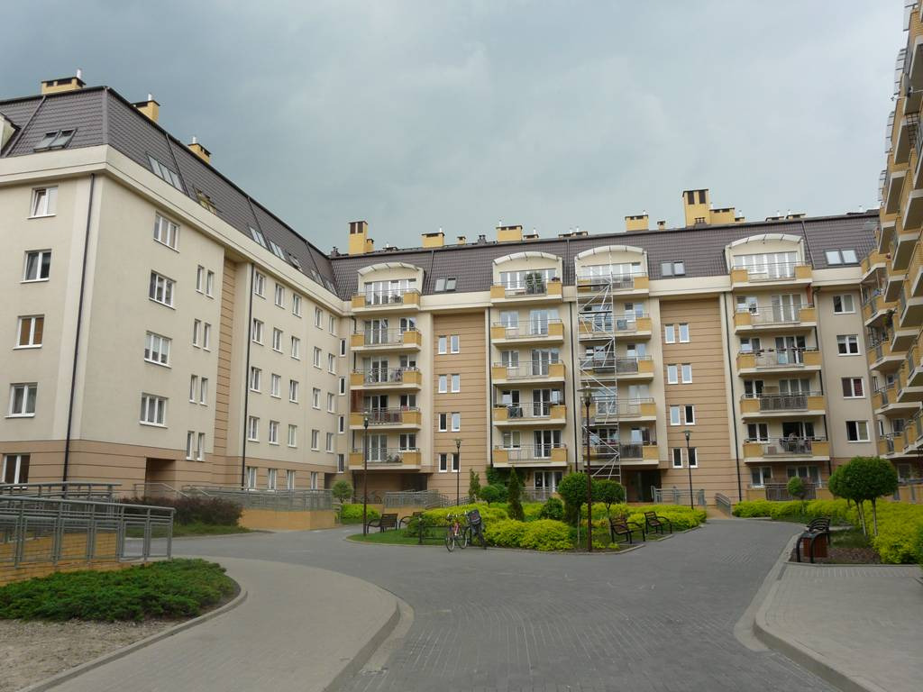 Kompleks budynków mieszkalnych z brązowym dachem wielospadowym i balkonami, widok z poziomu dziedzińca z kostki brukowej w pochmurny dzień.