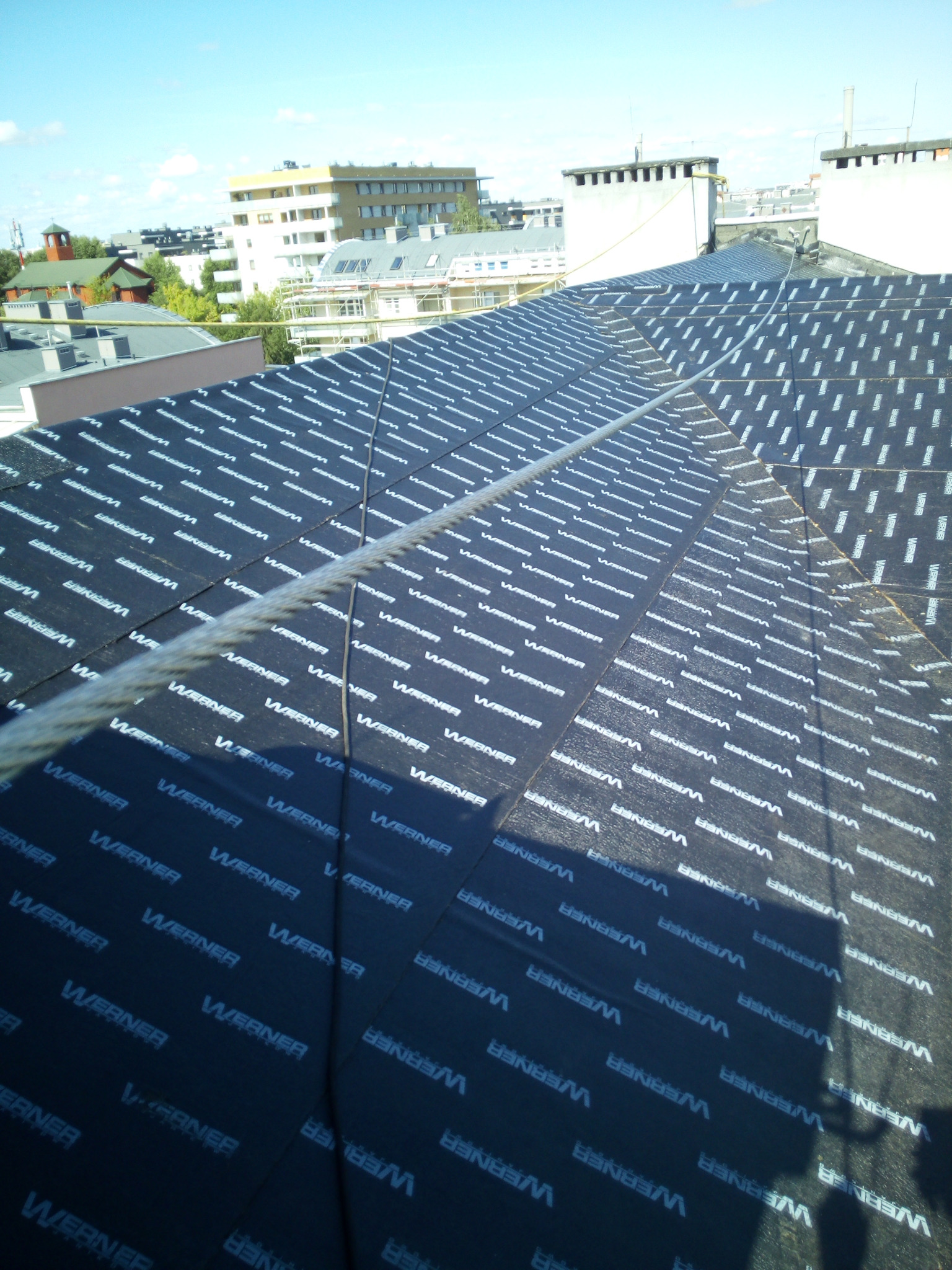 Czarna membrana dachowa z nadrukiem 'Werner' ułożona na dachu budynku mieszkalnego, widok z góry z linką zabezpieczającą przecinającą kadr.
