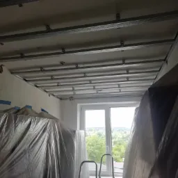 Widok na metalową konstrukcję podwieszanego sufitu w trakcie montażu, zabezpieczone folią meble i drabina przy oknie.