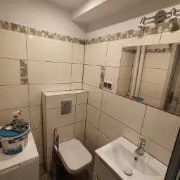 Umywalka z szafką i toaleta w małej łazience wykończonej jasnymi płytkami z dekoracyjnym pasem, widoczne akcesoria malarskie na pralce.