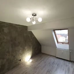 Pokój na poddaszu z szarą ścianą o fakturze betonu, drewnopodobną podłogą, grzejnikiem pod oknem dachowym i nowoczesną lampą sufitową.