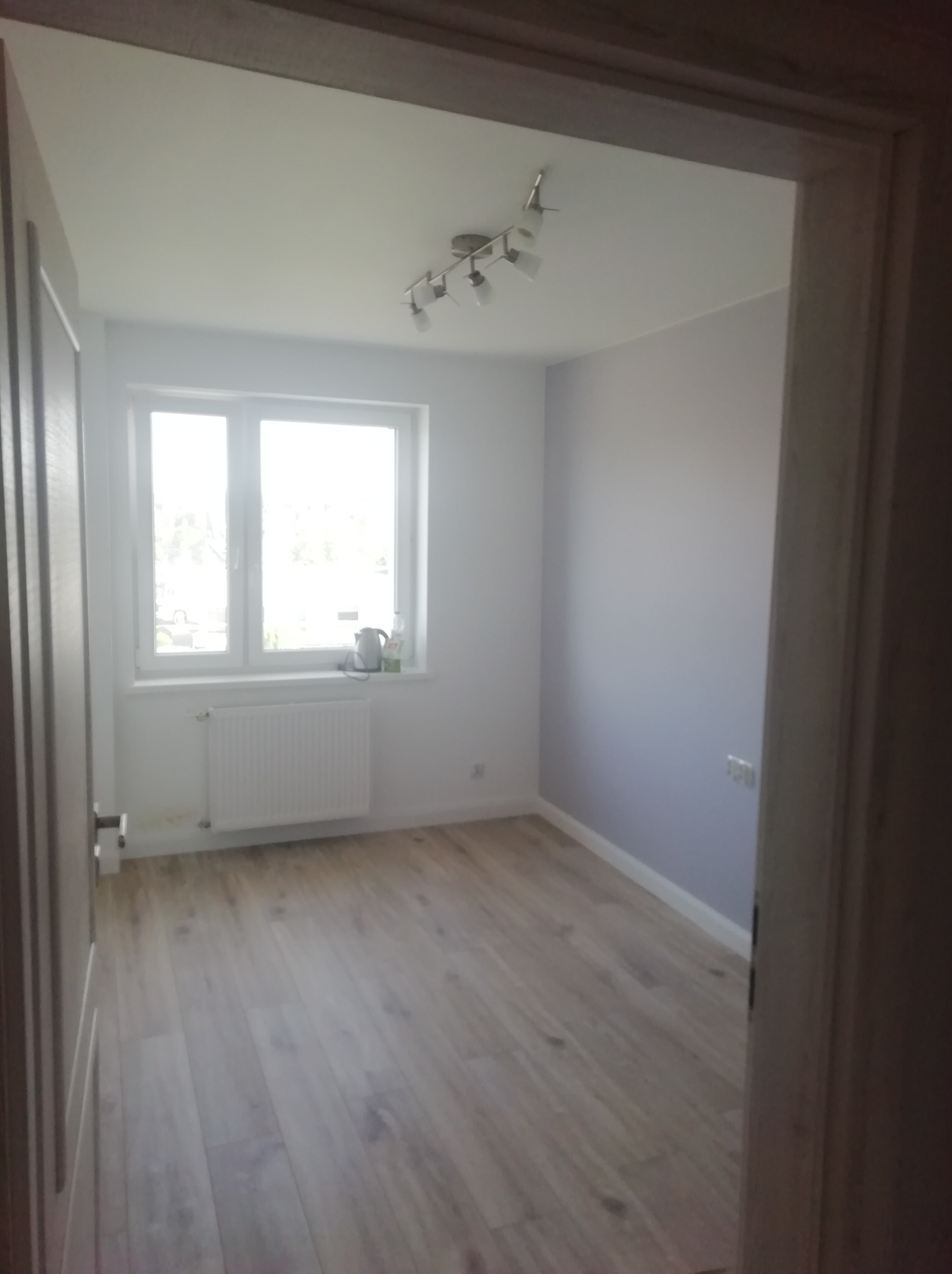 Pomalowane na szaro wnętrze pokoju w Miastku z jasną podłogą, grzejnikiem pod oknem i lampą sufitową