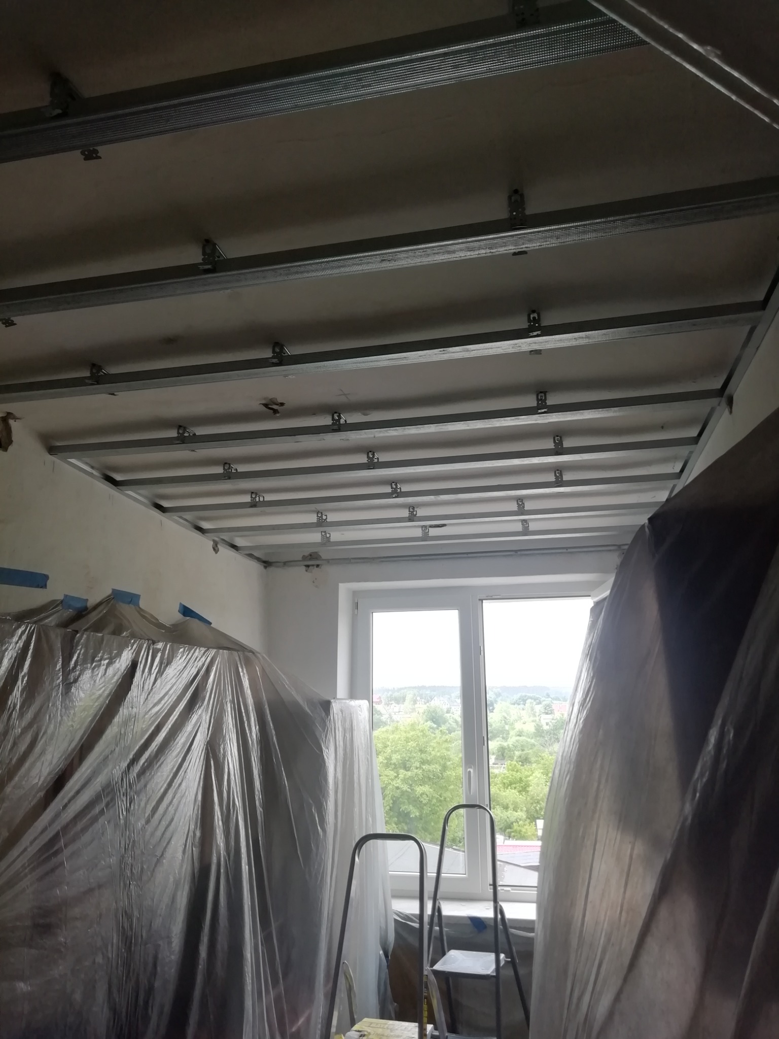 Widok na metalową konstrukcję podwieszanego sufitu w trakcie montażu, zabezpieczone folią meble i drabina przy oknie.