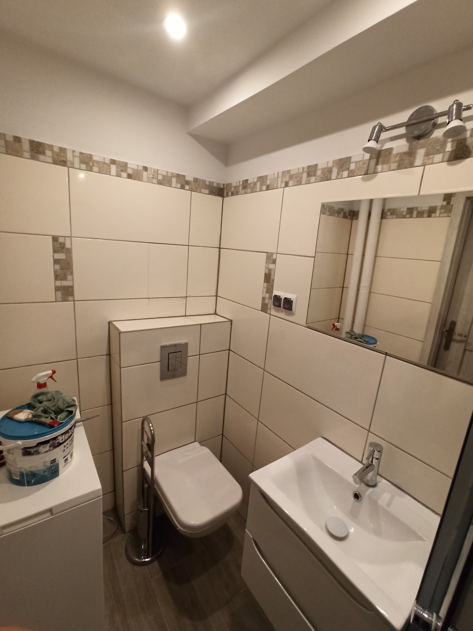 Umywalka z szafką i toaleta w małej łazience wykończonej jasnymi płytkami z dekoracyjnym pasem, widoczne akcesoria malarskie na pralce.