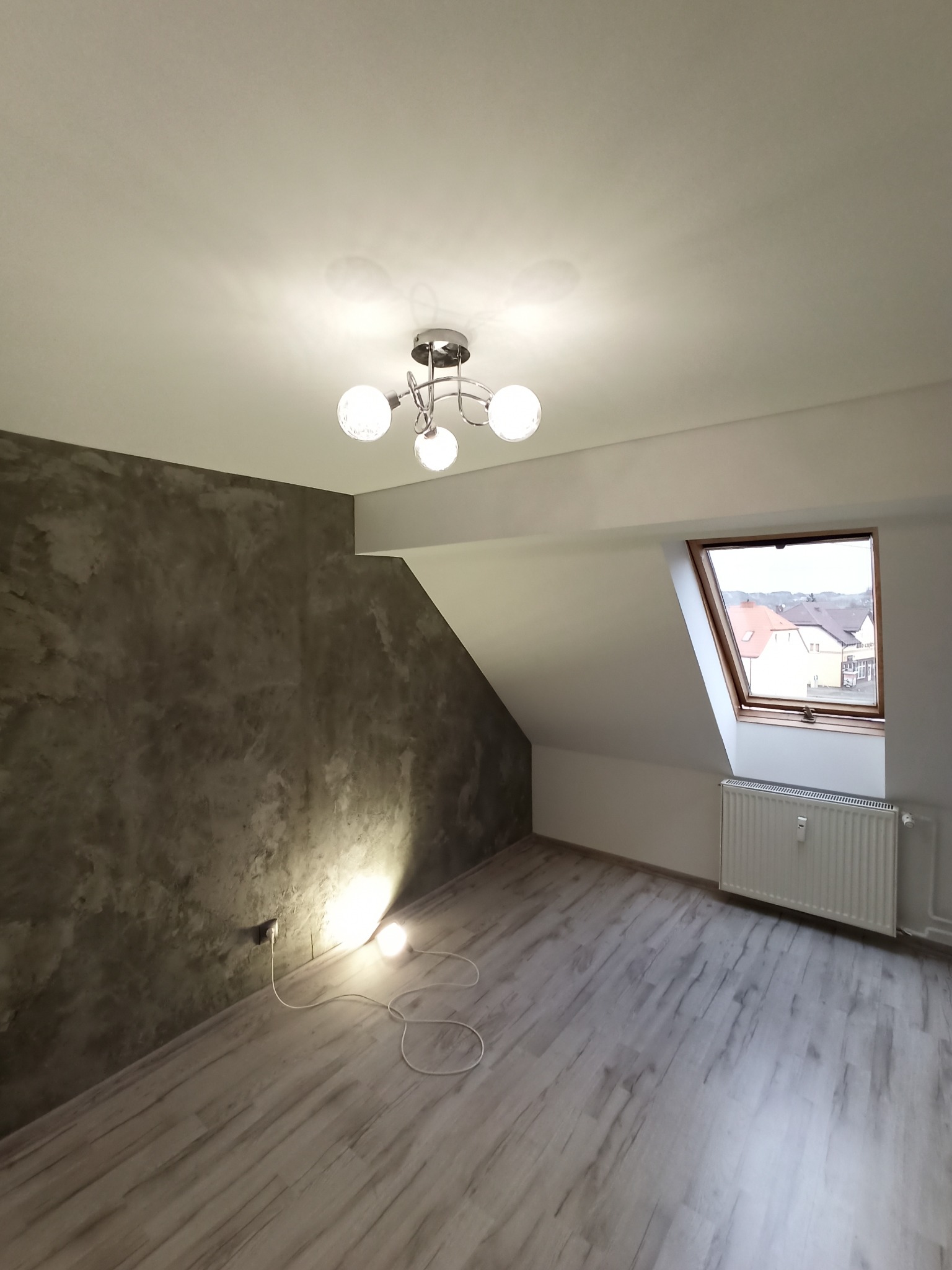 Pokój na poddaszu z szarą ścianą o fakturze betonu, drewnopodobną podłogą, grzejnikiem pod oknem dachowym i nowoczesną lampą sufitową.