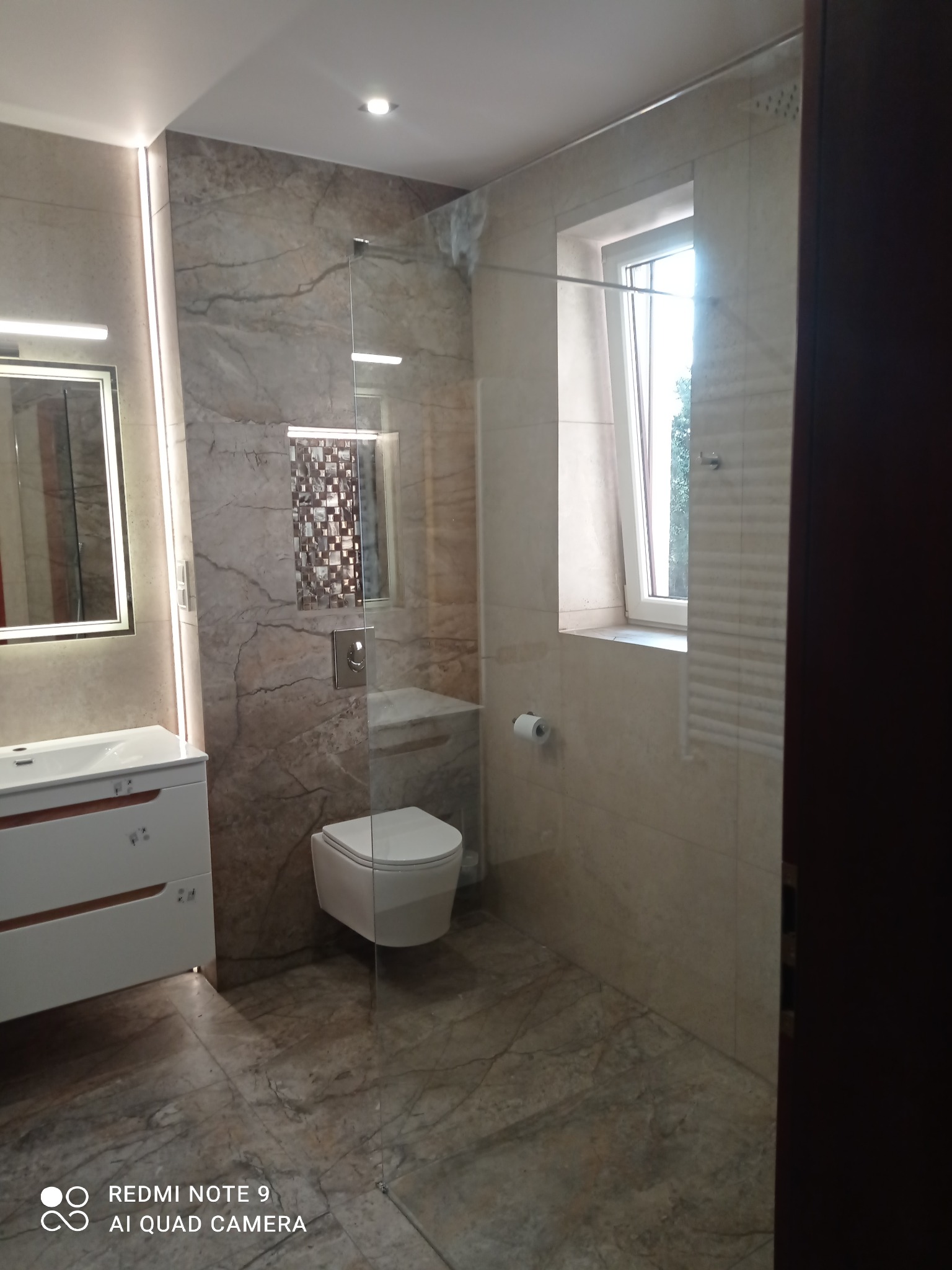 Nowoczesna łazienka z kabiną prysznicową typu walk-in, marmurowymi płytkami i podświetlanym lustrem. Uwagę zwraca mozaika w strefie prysznica i minimalistyczna armatura.