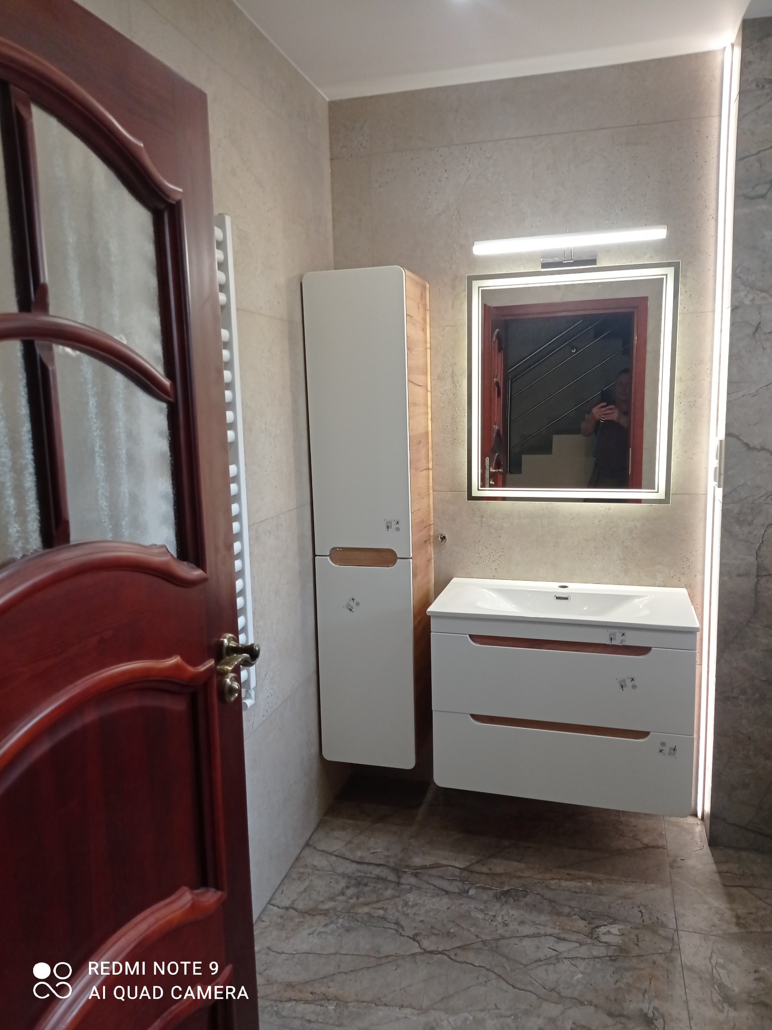 Ukończona łazienka z szafką pod umywalką, wysoką szafką i lustrem z oświetleniem LED. Widoczna część otwartych drzwi z ciemnego drewna.