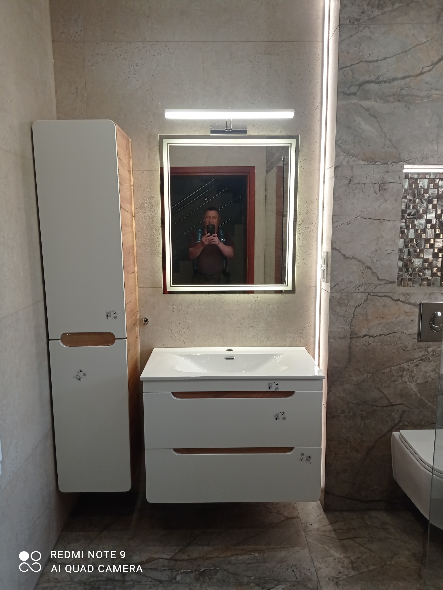 Nowoczesna łazienka z podświetlanym lustrem, szafką pod umywalką i wysoką szafką boczną. Na ścianach beżowe płytki imitujące marmur, mozaika dekoracyjna.